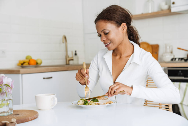 Молода чорна жінка посміхається, обідаючи на кухні вдома
 - Фото, зображення