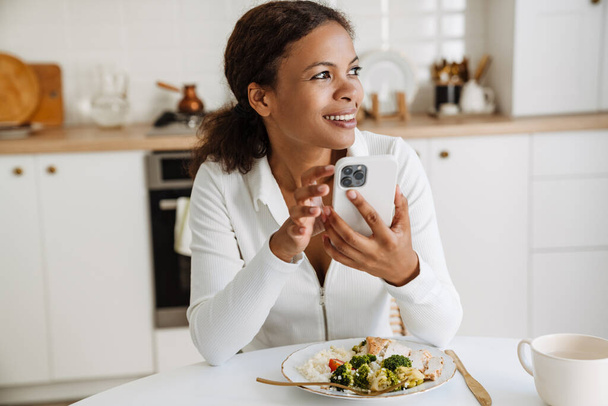 Black woman smiling and using cellphone while having lunch at home - Foto, imagen