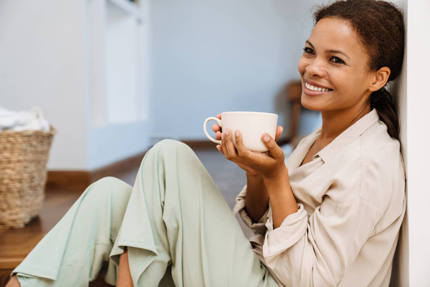 Zwarte vrouw glimlachen en het drinken van thee terwijl zitten op de trap thuis - Foto, afbeelding