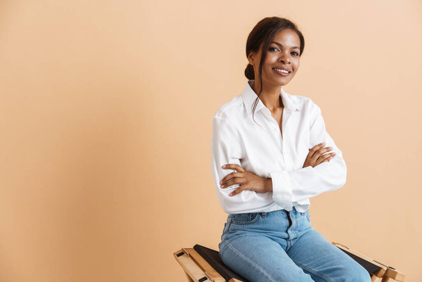 Young black woman wearing shirt smiling while sitting on chair isolated over beige background - Foto, Bild