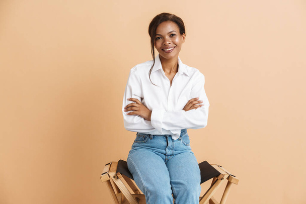 Young black woman wearing shirt smiling while sitting on chair isolated over beige background - Foto, imagen