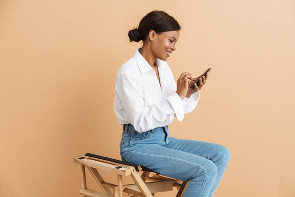 Молода чорна жінка використовує мобільний телефон, сидячи на стільці ізольовано на бежевому фоні
 - Фото, зображення