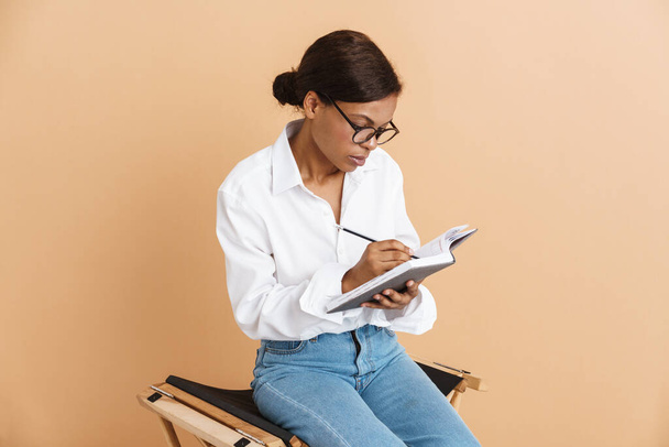 Young black woman writing down notes while sitting on chair isolated over beige background - 写真・画像