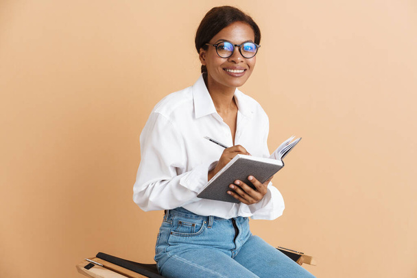 Young black woman writing down notes while sitting on chair isolated over beige background - Foto, immagini