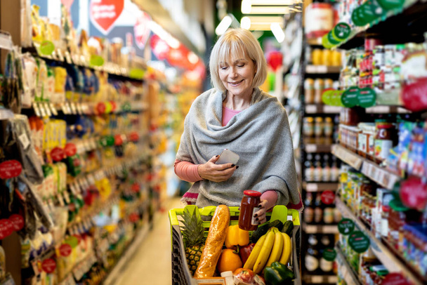 Vanhempi nainen asiakas käyttää matkapuhelinta ostoksia supermarketissa - Valokuva, kuva