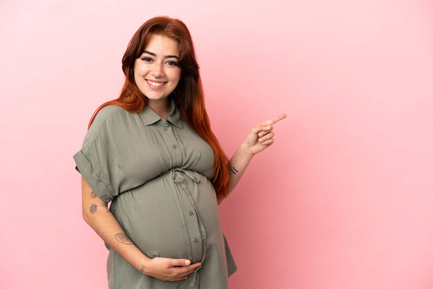 Young redhead caucasian woman isolated on pink background pregnant and pointing to the lateral - Foto, Imagem
