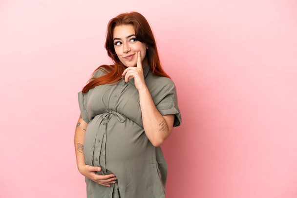 Joven pelirroja caucásica aislada sobre fondo rosa embarazada y pensando - Foto, Imagen