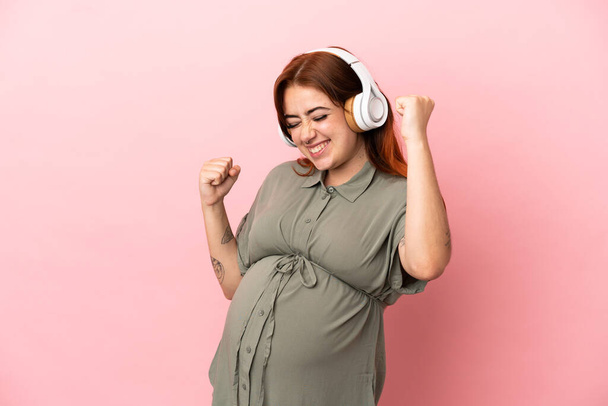 Jeune rousse caucasienne isolée sur fond rose enceinte et écoutant de la musique - Photo, image