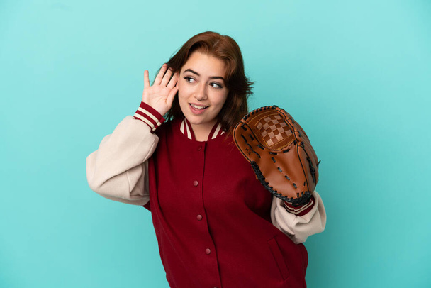 Mladý ryšavý žena hrát baseball izolovaný na modrém pozadí poslech něco tím, že ruku na ucho - Fotografie, Obrázek