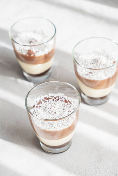 Тришаровий шоколад і ванільний десерт мусу в скляних чашках, посипаний кокосовими пластівцями, на дитячій дошці. брошура
 - Фото, зображення