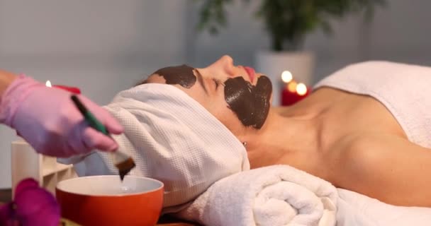 Cosmetólogo aplica máscara negra rejuvenecedora en la cara de la mujer en el spa - Imágenes, Vídeo