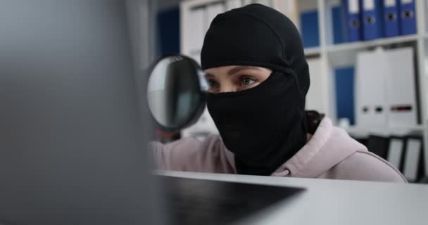 Ληστής που φοράει μάσκα και ψάχνει πληροφορίες χρησιμοποιώντας μεγεθυντικό φακό σε υπολογιστή στο γραφείο - Πλάνα, βίντεο