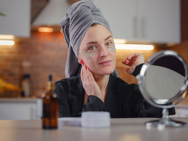 Patches for eyes. Attractive woman in kitchen with towel on her head glues transparent patches to skin under her eyes. Hygiene, beautician, makeup, face care concept. - Foto, Bild