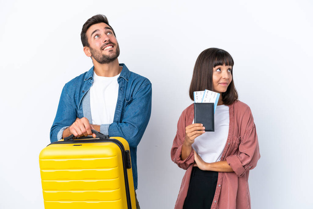 Young traveler couple holding a suitcase and passport isolated on white background looking up while smiling - Foto, afbeelding