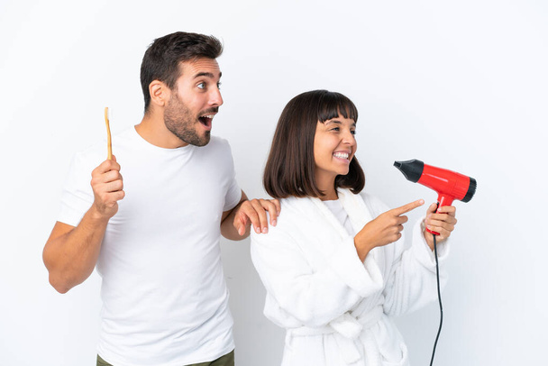 Young caucasian couple holding a hairdryer and toothbrush isolated on white background pointing to the side to present a product - Foto, afbeelding