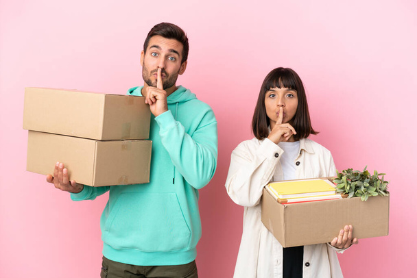 mladý kráska pár pohybující se v novém domově mezi krabicemi izolované na růžovém pozadí ukazující znamení mlčení gesto dát prst do úst - Fotografie, Obrázek