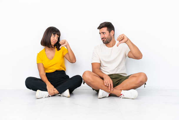 Young couple sitting on the floor isolated on white background showing thumb down sign with negative expression - Foto, Bild