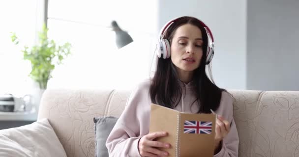 Woman in headphones is studying English at home sitting on couch - Materiał filmowy, wideo