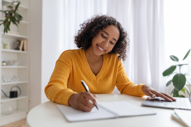 Mujer negra joven que estudia o trabaja en línea con el ordenador portátil en casa, tomando notas, disfrutando de trabajo a distancia o educación - Foto, imagen