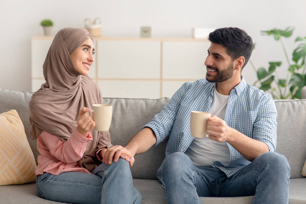 feliz joven musulmán pareja tener café sentado en sofá interior - Foto, imagen