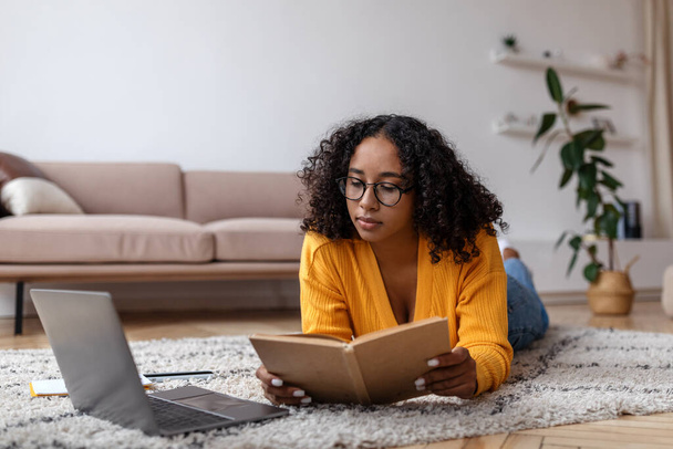 Keskittynyt nuori musta nainen käsittelyssä kirja, makaa lattialla lähellä kannettavaa, opiskelee verkossa kotoa - Valokuva, kuva
