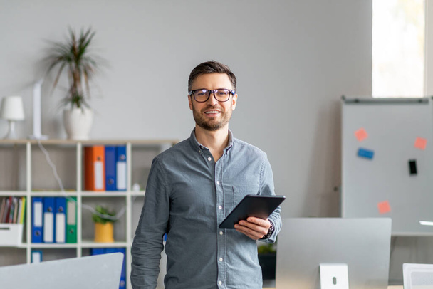 Портрет щасливого зрілого офісного працівника, який тримає цифровий планшет, дивлячись і посміхаючись на камеру, стоїть біля робочого місця
 - Фото, зображення