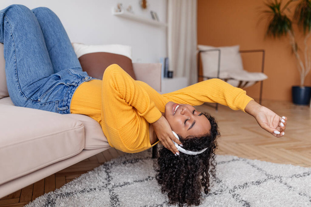 Fröhliche junge schwarze Frau mit Kopfhörern liegt mit geschlossenen Augen auf dem Sofa und hört zu Hause Musik - Foto, Bild