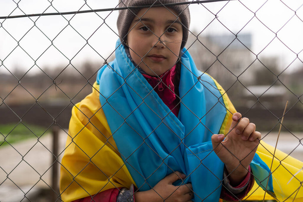 sad little girl with the flag of Ukraine behind a metal fence. Social problem of refugees and forced migrants - Foto, imagen