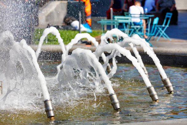 Αφρίζοντες πίδακες νερού από ένα συντριβάνι της πόλης με πιτσιλιές στο φως του ήλιου με φόντο ένα καλοκαιρινό καφέ στη θολούρα. - Φωτογραφία, εικόνα