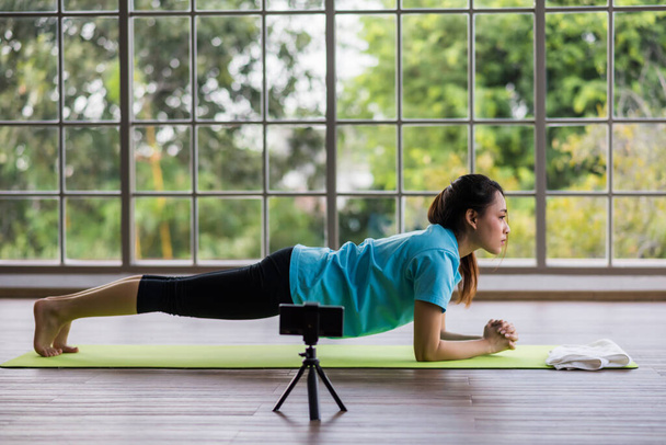 Young Asian yoga trainer recording video content for her online exercise course with mobile phone and tripod. She show planking to smartphone to live streaming. - Photo, image