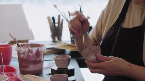 Μια ηλικιωμένη γυναίκα ζωγραφίζει ένα μικρό πήλινο κύπελλο με σκονισμένο χρώμα τριαντάφυλλου - Πλάνα, βίντεο