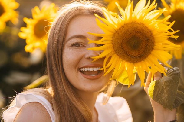 a cheerful girl with freckles holds a sunflower and covers her face against the background of a sunset and a field. - Φωτογραφία, εικόνα