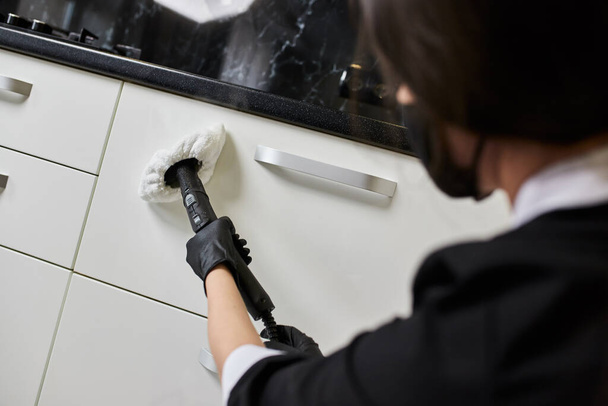 Ο υπάλληλος της εταιρείας καθαρισμού με λαστιχένια γάντια απομακρύνει τη βρωμιά από την επιφάνεια εργασίας χρησιμοποιώντας επαγγελματική ατμοκαθαρίστρια στην κουζίνα, απολύμανση και αποστείρωση - Φωτογραφία, εικόνα
