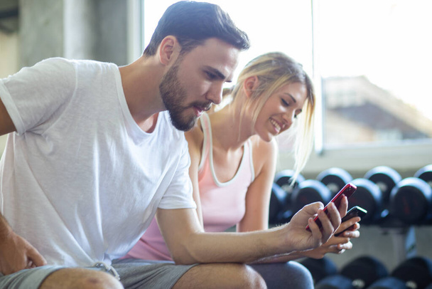 Couple of men women use smartphone talking together after working out in fitness gym - Photo, Image