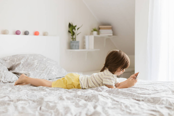 Un petit garçon de quatre ans se couche sur le lit et joue un téléphone portable, un smartphone. Les enfants utilisent la technologie. Le problème des enfants et des gadgets - Photo, image