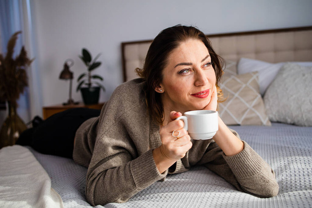Successful middle aged woman smiling at home. A calm and peaceful middle-aged woman takes a break, rests with a cup of hot tea on bed at home. Contented charming mature lady enjoying coffee.  - Foto, afbeelding