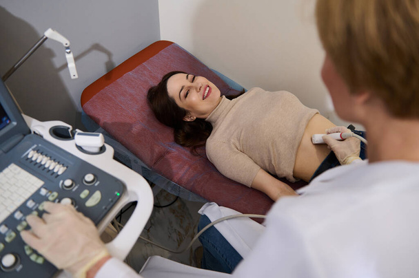 Encantadora mujer caucásica sonriendo a su médico asistente mientras revisa su salud durante un examen de ultrasonido en una clínica médica. ultrasonografía USG - Foto, imagen