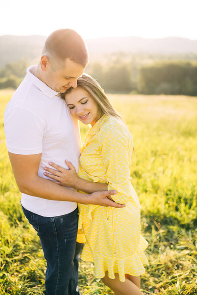 Ένας νεαρός σύζυγος και η έγκυος γυναίκα του αγκαλιάζονται σε ένα ηλιόλουστο καλοκαιρινό χωράφι. - Φωτογραφία, εικόνα