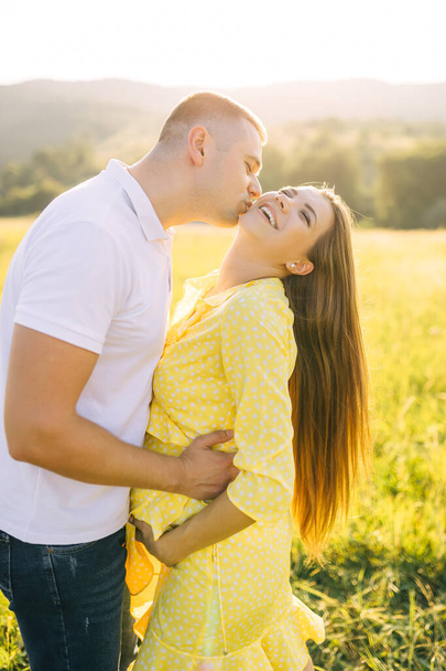 young beautiful couple girl pregnant in yellow dress and man in white t-shirt kissing in sunny field - Фото, изображение