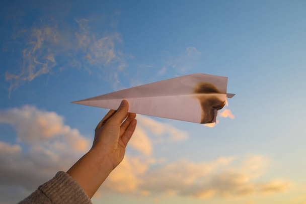 A burning paper airplane in the background of the sky at sunset. A hand holds a burning paper airplane. - Photo, Image