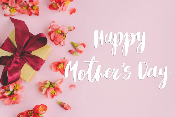 Χαρούμενη ευχετήρια κάρτα της μητέρας. Ευτυχισμένες μητέρες ημέρα κείμενο και κουτί δώρου με κόκκινα λουλούδια σε ροζ φόντο επίπεδη θέσει. Κομψή κάρτα λουλουδιών. Χειρόγραφα γράμματα. Ημέρα μητέρων - Φωτογραφία, εικόνα