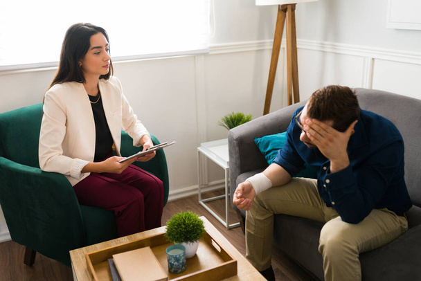 Przygnębiony biały mężczyzna z zabandażowanym nadgarstkiem rozmawiający z terapeutą o swoich problemach z pomocą psychiczną - Zdjęcie, obraz