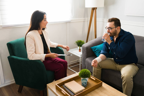 Beautiful female psychologist giving a glass of water to a man with depression. Patient talking to the therapist about the suicide attempt - Foto, imagen