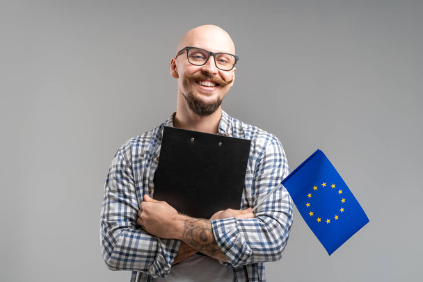 Millennial caucasian businessman holding clipboard and European Union flag. Young balded bearded guy. - Foto, afbeelding