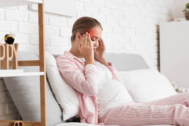 Mladá těhotná žena s bolestí hlavy sedí doma na posteli - Fotografie, Obrázek