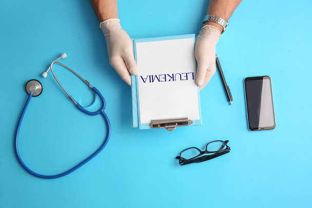 Manos del médico sujetando el portapapeles con la palabra LEUKEMIA, estetoscopio, teléfono y gafas sobre fondo azul, vista superior - Foto, Imagen