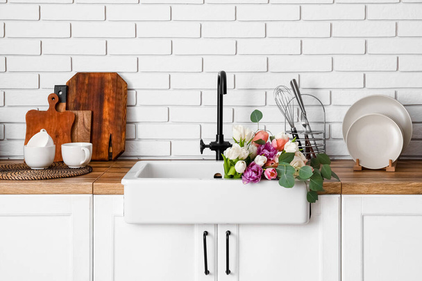 Évier avec de belles fleurs et ustensiles de cuisine sur les comptoirs près du mur de briques blanches - Photo, image
