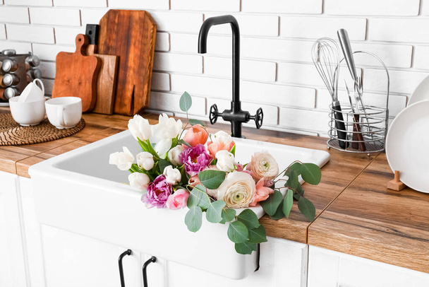 Spüle mit schönen Blumen und Küchenutensilien auf Theken in der Nähe der weißen Ziegelwand - Foto, Bild