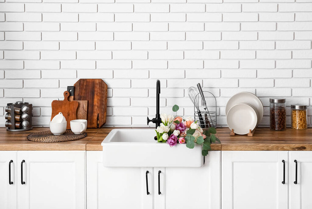 Βουτιά με όμορφα λουλούδια και σκεύη κουζίνας σε πάγκους κοντά σε λευκό τοίχο από τούβλα - Φωτογραφία, εικόνα