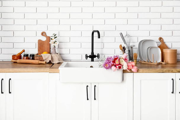 Ceramiczne zlewozmywak z pięknymi kwiatami i naczynia kuchenne na licznikach w pobliżu ściany z białej cegły - Zdjęcie, obraz